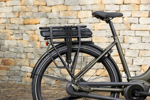 Zijkant van een e-bike met een bagagedrager en accu, geplaatst voor een stenen muur.